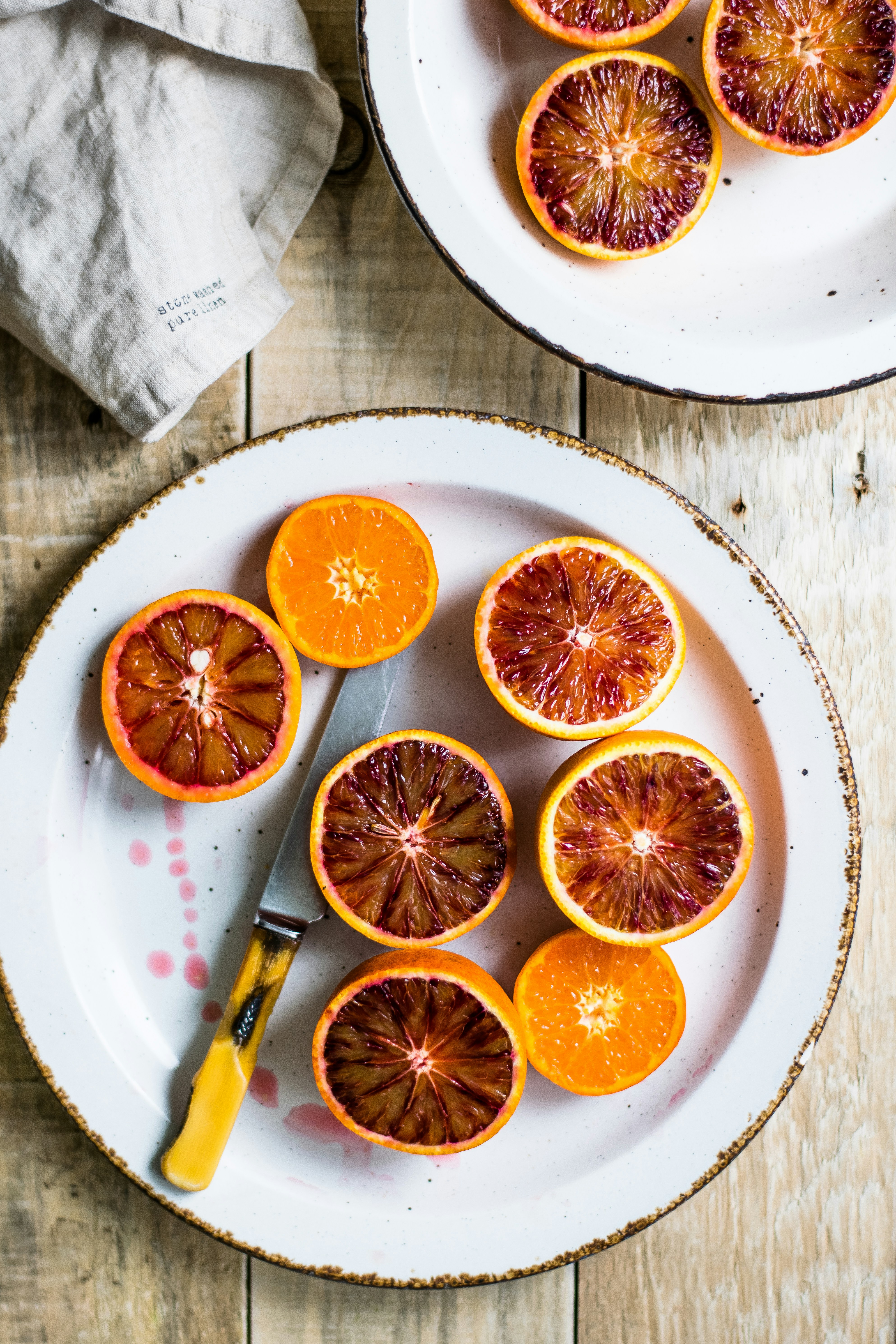Halved blood oranges arranged on white plates with a small knife, showing varied red and orange interiors on a wooden surface.